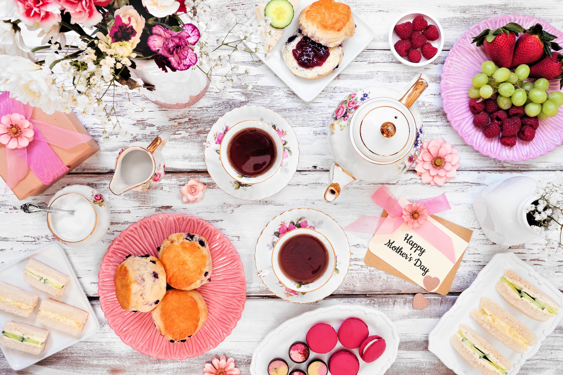 Mother's Day tea table scene over a white wood background. Greeting card, gift, flowers, vintage tea set, finger sandwiches, chocolates, desserts and fruit.